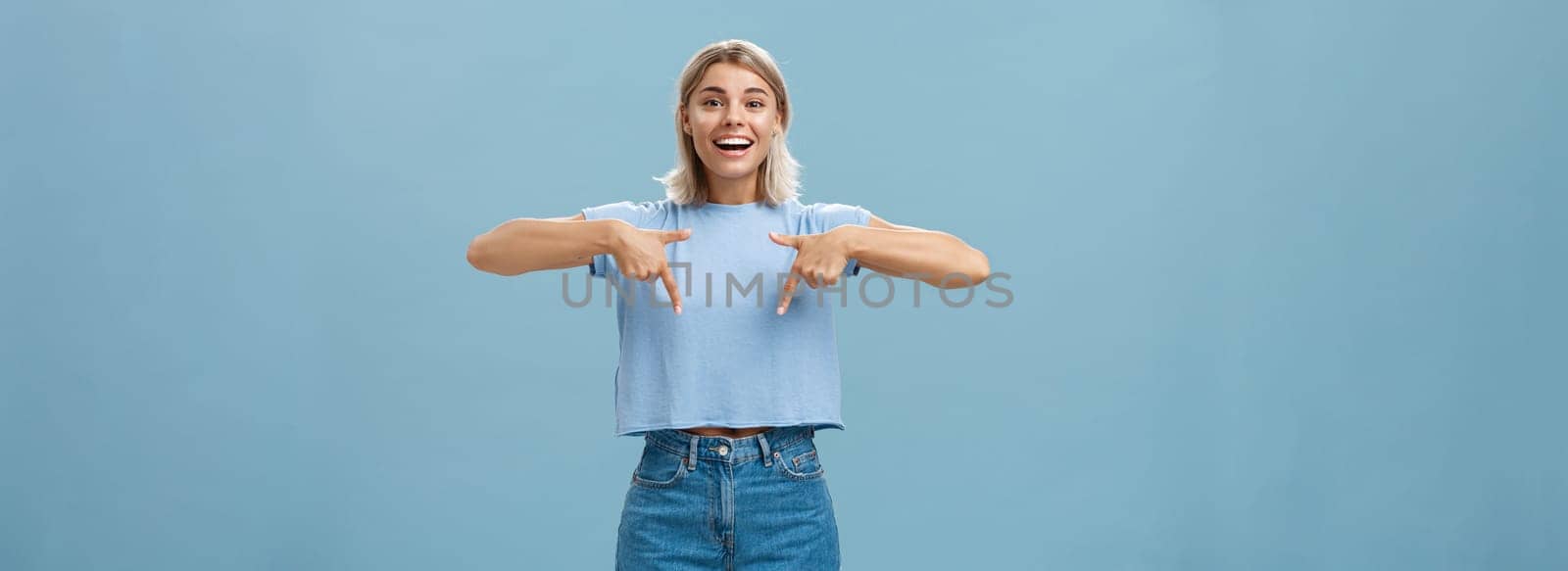 You have to see this. Portrait of joyful amused and happy stylish blonde female in trendy t-shirt pointing down with arms near chest smiling broadly showing amazing copy space over blue background.