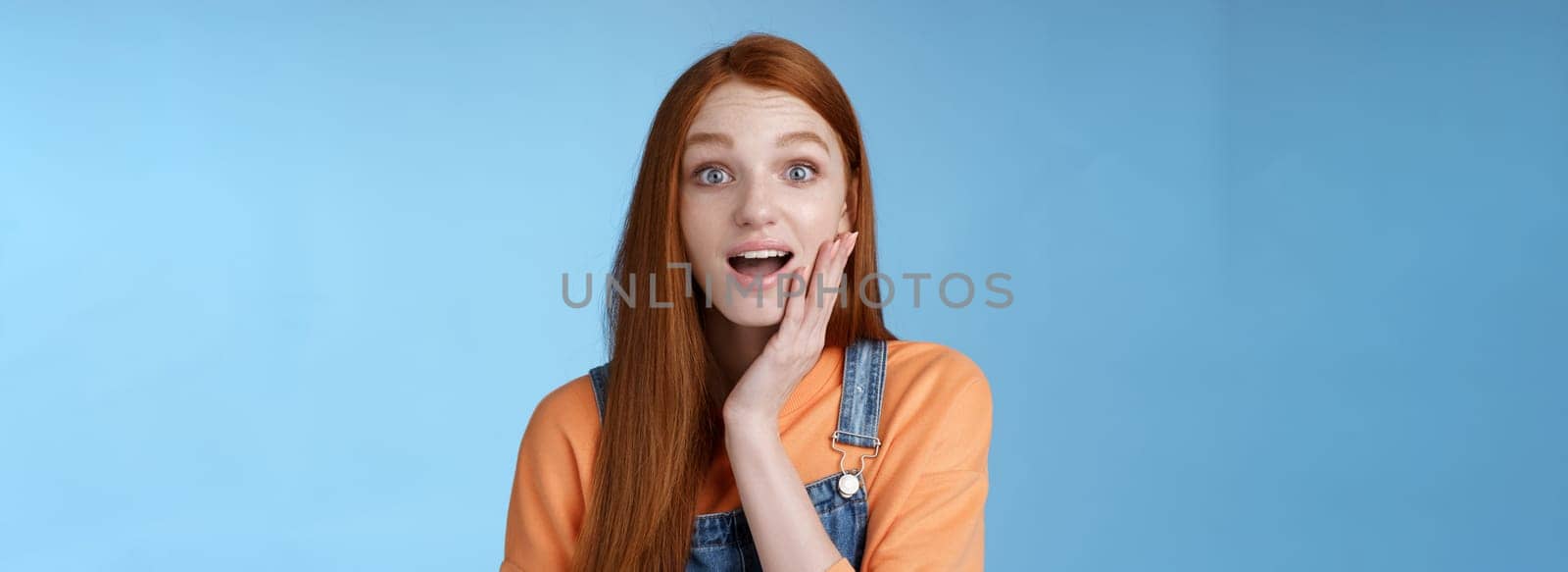 Amazed friendly sensitive impressed redhead girl learn awesome pleasant news open mouth wondered astonished smiling glad friend touch cheek amused, standing blue background happy. Copy space