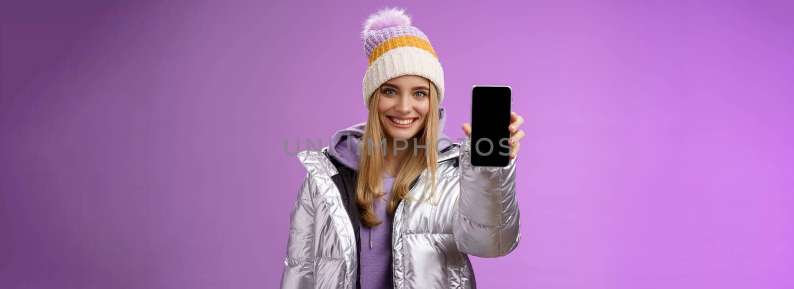 Friendly cheerful confident blond girl in silver stylish winter jacket hat extend arm showing smartphone display advertising awesome new device app smiling self-assured recommend use mobile phone by Benzoix
