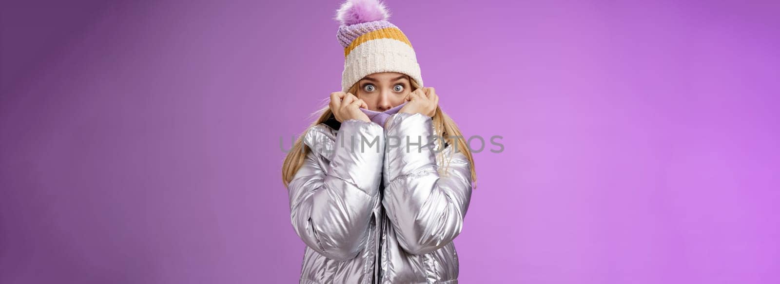 Afraid shocked young charming sister pulling jacket face cover hiding frightened scared terrifying stories snowman walking mountains widen eyes concerned look camera fear, standing purple background.