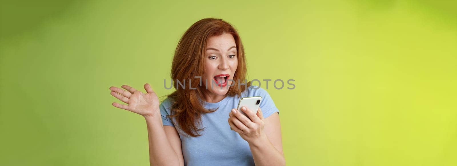 Surprised glad middle-aged pretty redhead woman look smartphone raise hand celebratory excitement open mouth reacting impressed gladly read good excellent news look mobile screen green background by Benzoix
