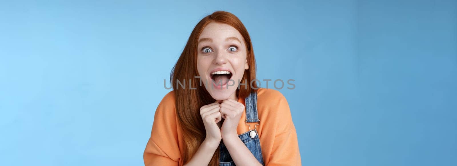 Cute redhead european girl blue eyes freckles reacting amused shocking rumor lift eyebrows drop jaw surprised smiling excited picked get role theatre play rejoicing astonished blue background by Benzoix