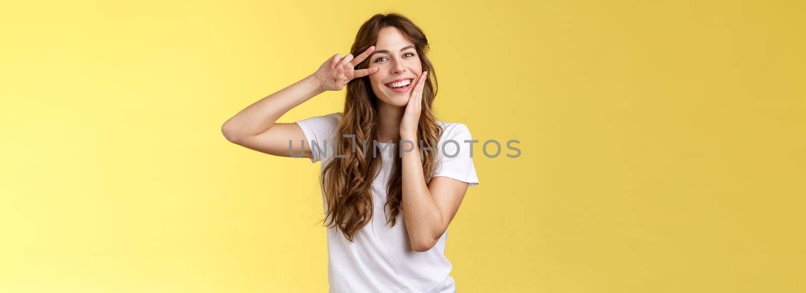 Cheerful positive lovely girlfriend curly hairstyle touch cheek blushing modest cute showing victory peace sign near eye optimistic upbeat attitude having fun enjoying summer yellow background. Lifestyle.