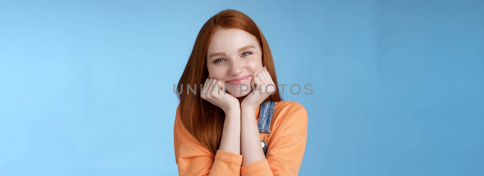 Tenderness, wellbeing, beauty conept. Attractive young ginger girl pure skin blue eyes lean palms silly smiling camera look amused enthusiastic listen interesting stories gladly, blue background.