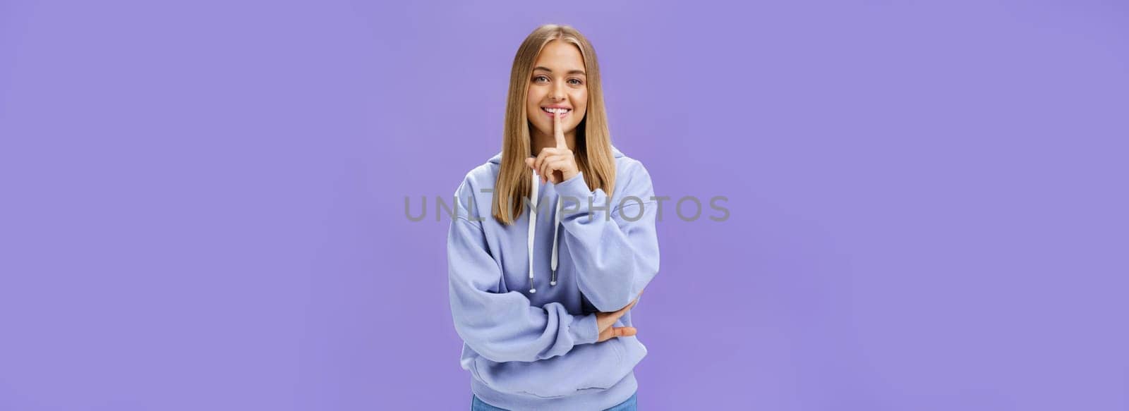 Portrait of joyful cute feminine young woman in hoodie smiling happily showing shush gesture hiding surprise asking keep secret standing amused and carefree against purple background. Copy space