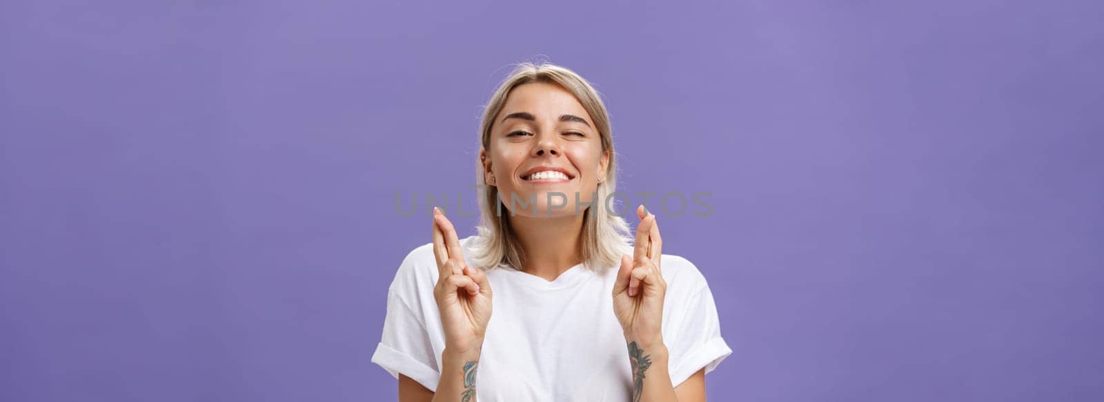 Waist-up shot of hopeful optimistic attractive stylish woman in white t-shirt with tattooed arms winking smiling joyfully while crossing fingers for good luck making wish over purple background by Benzoix