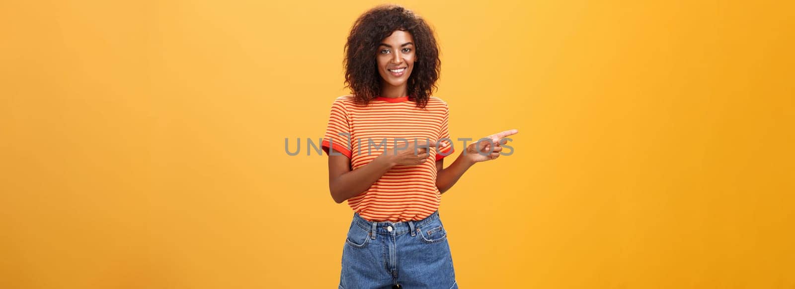 Portrait of charming friendly-looking african-american female coworker in casual outfit pointing left and smiling broadly showing perfect copy space for customers against orange background. Lifestyle.