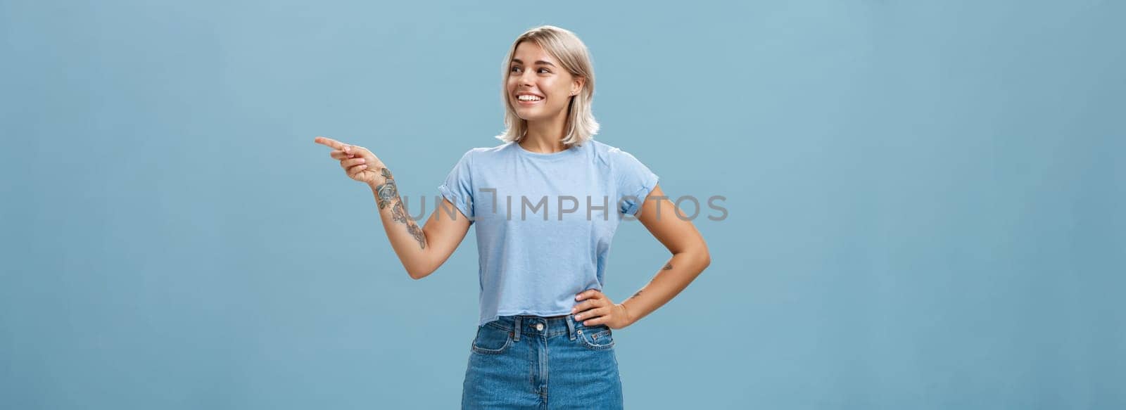 Bossy confident female manager with tattoo on arm holding hand on waist pointing and gazing right with pleased relaxed look giving directions to employees posing over blue background by Benzoix