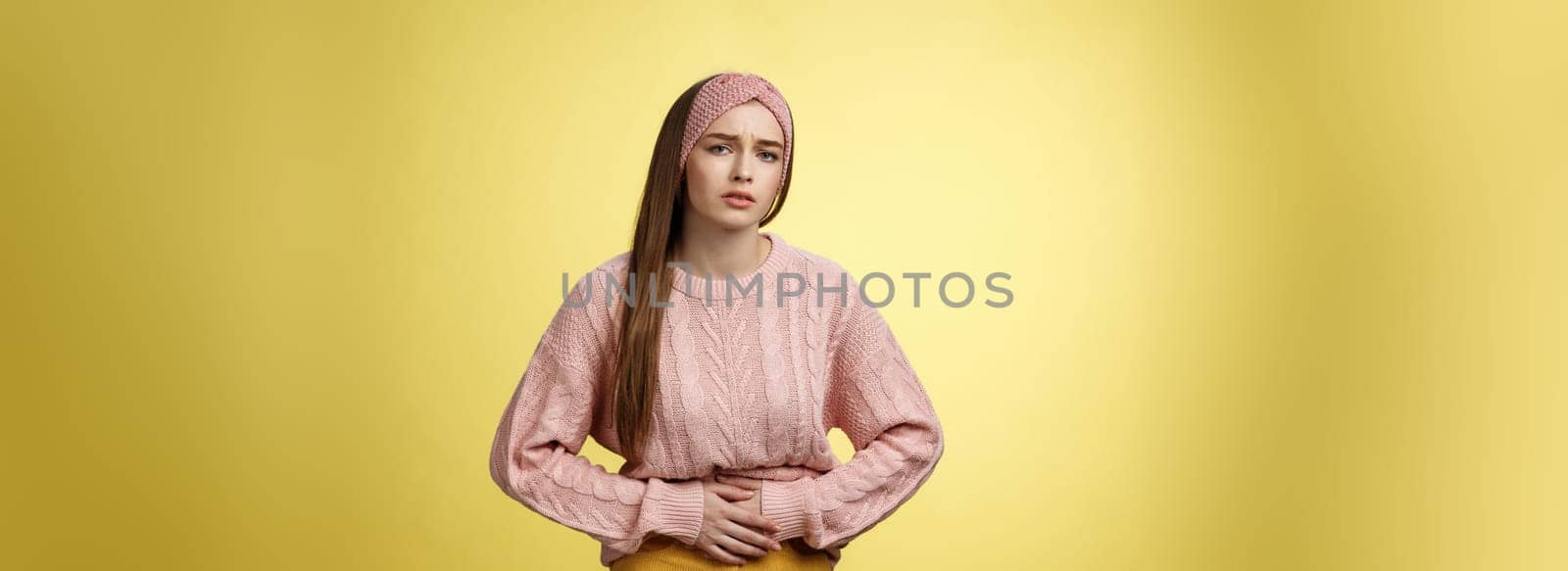 Girl suffering pain in stomach, having painful periods, feeling unwell touching belly grimacing displeased and troubled, having cramps, stooping upset of stomachahe against yellow background.