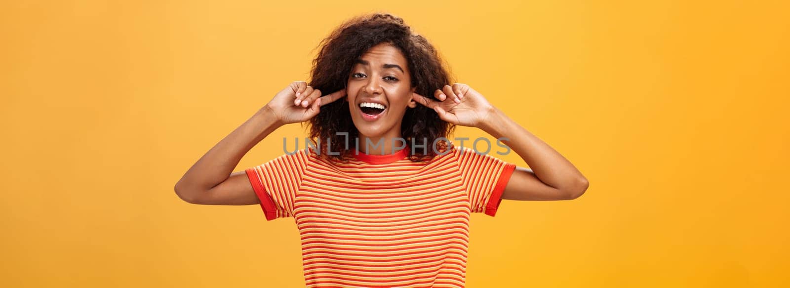 Do not care what saying cannot hear you. Carefree indifferent good-looking african american woman with curly hairstyle closing ears with index finger singing lalala during argument behaving childish by Benzoix