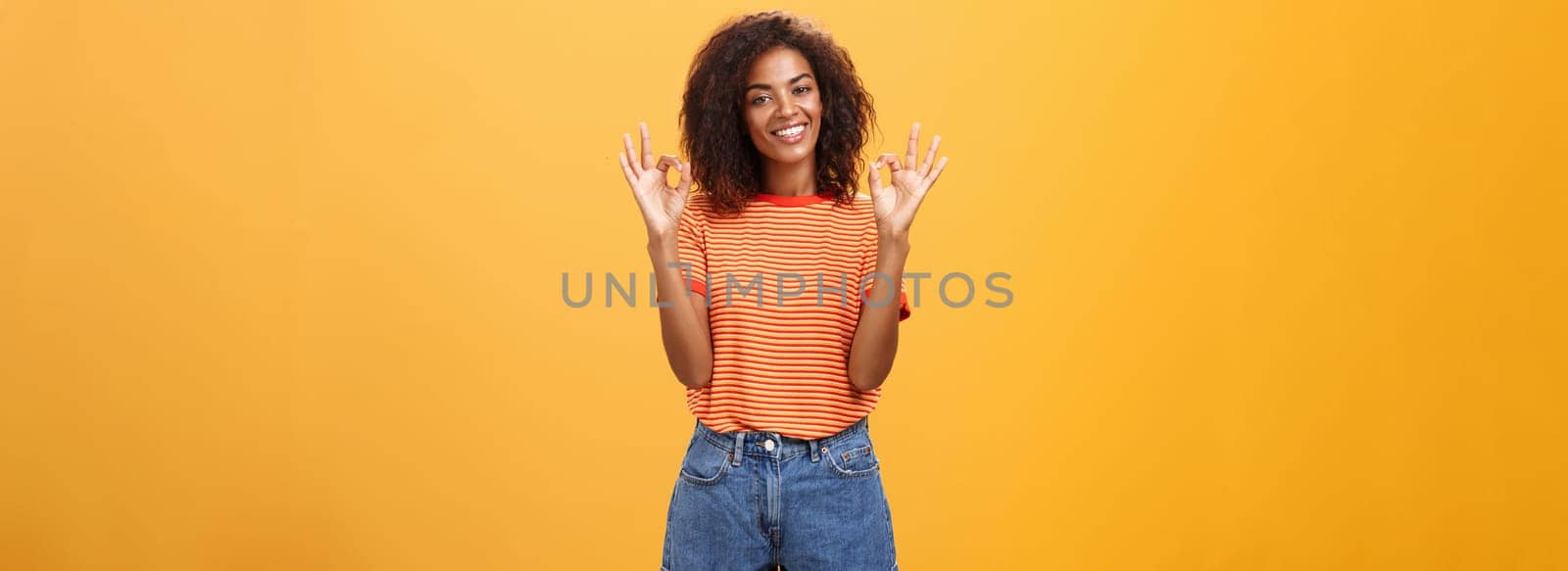 Chill attractive satisfied african american female customer picking new outfit being delighted and pleased with cool staff raising hands in okay or awesome gesture smiling happily over orange wall by Benzoix