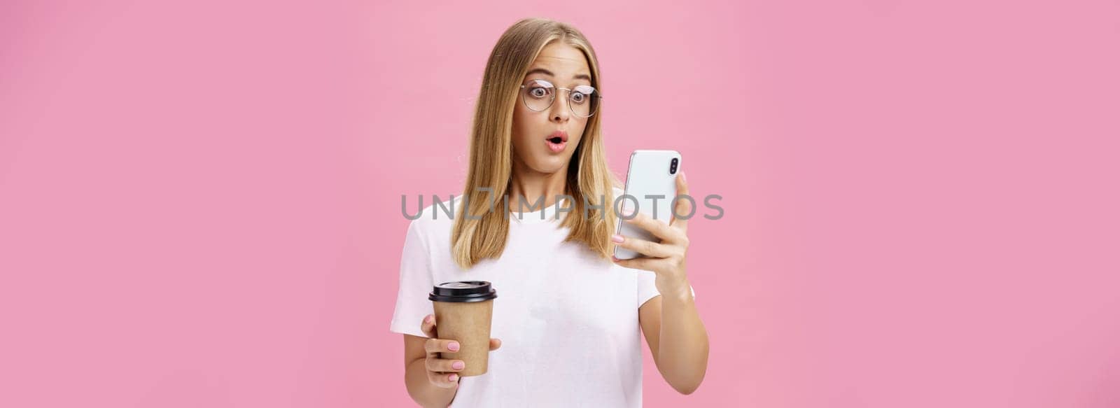 Woman drinking coffee being shocked by received message reacting on stunning news folding lips gasping looking astonished and impressed at smartphone screen holding paper cup, posing over pink wall. Technology concept