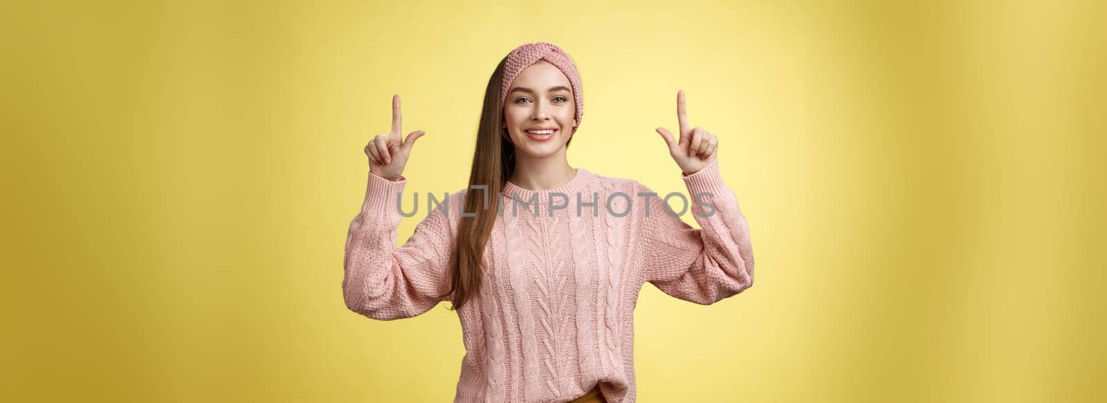 Easygoing beautiful young female student in knitted sweater, headband pointing up, promoting advertisement, smiling happily, feeling positive posing in good mood, grinning at camera over yellow wall.