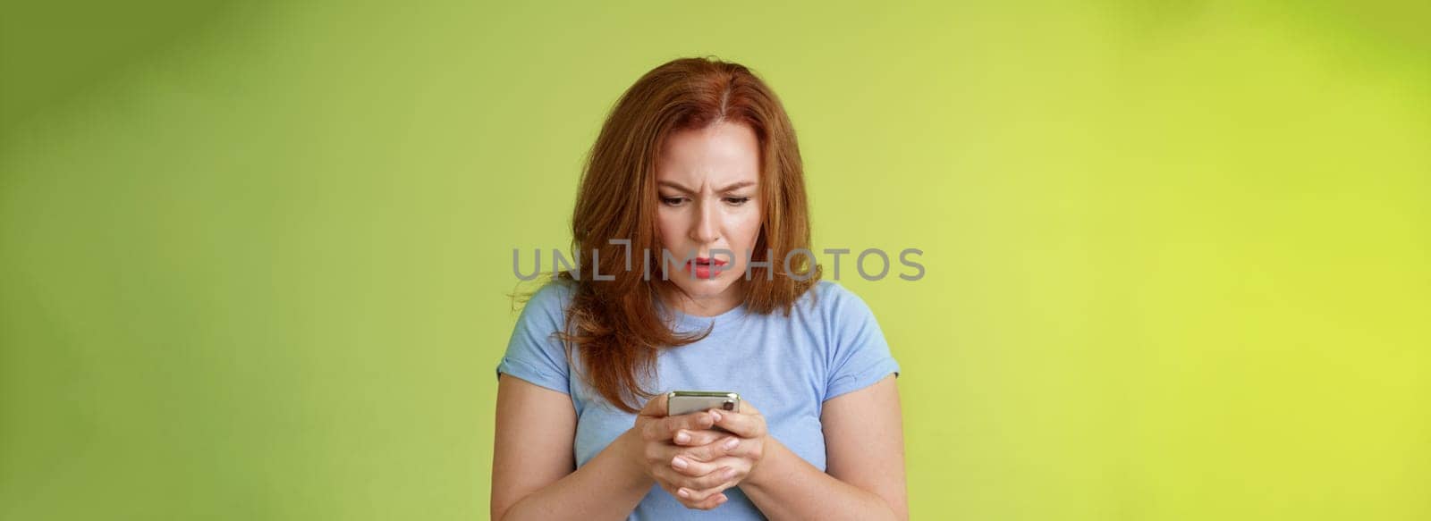 Confused unsure redhead middle-aged woman learn how use social media trying understand emoji look intense focused smartphone display reading important news message stand green background by Benzoix