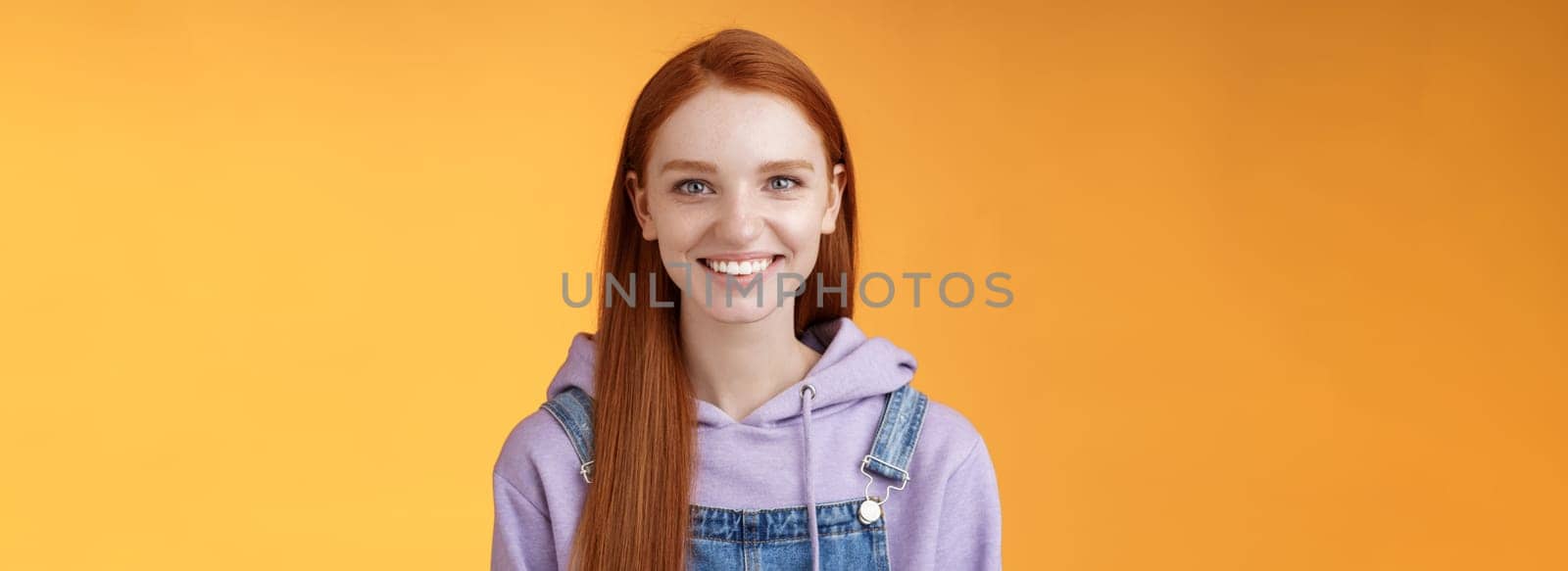 Attractive modern hipster young redhead girl smiling delighted relaxed talking have casual joyful day downtown walking friends wearing purple hoodie overalls, summer vibes, standing orange wall.