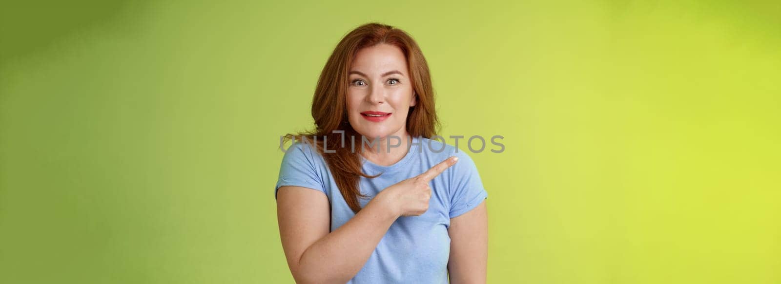 Surprised excited middle-aged wondered redhead woman pointing left amused standing thrilled joyful green background look camera curious interested cannot wait check-out great promo.