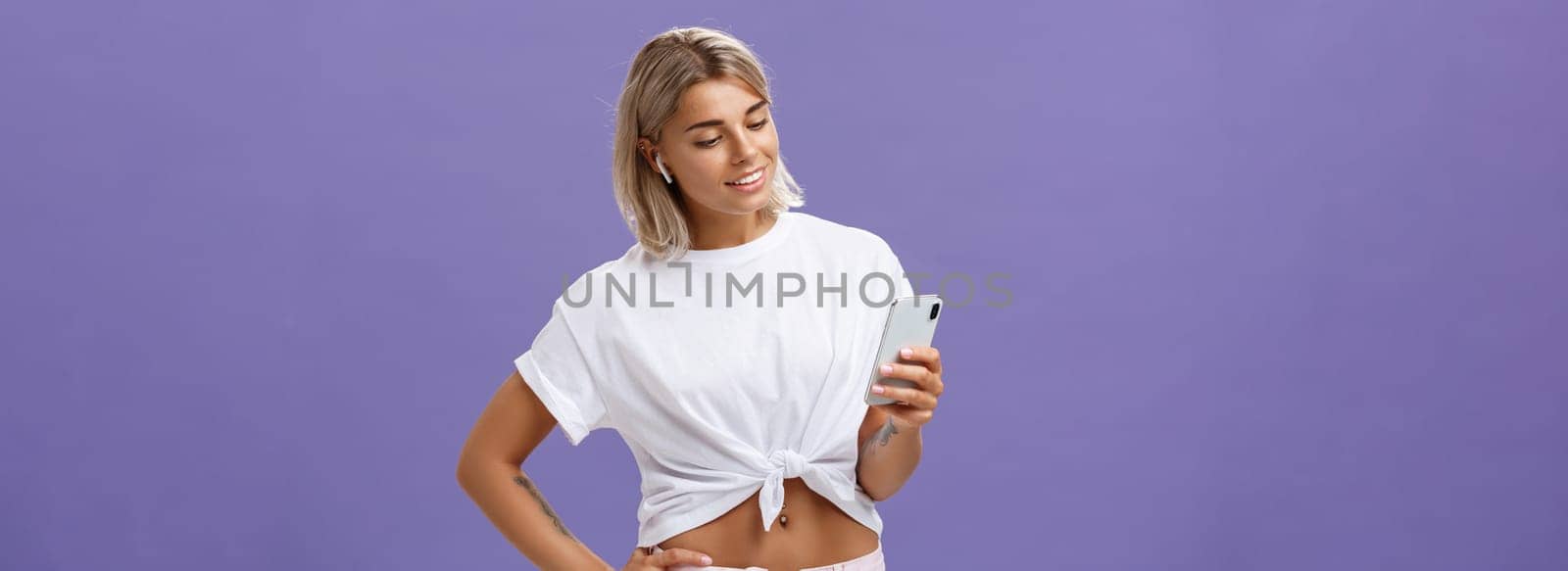 Checking out new track from favorite singer. Good-looking sociable and carefree european woman with tanned skin listening music in wireless earbuds picking song in smartphone over purple wall by Benzoix