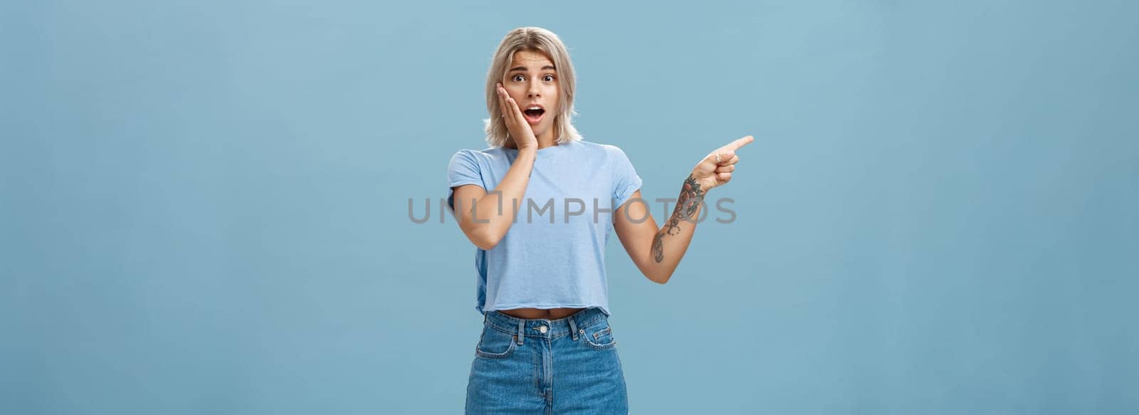 Lifestyle. Waist-up shot of worried surprised emotive blonde woman in t-shirt saying gosh opening mouth from shock holding palm on cheek looking with empathy while pointing left with tattooed arm over blue wall.