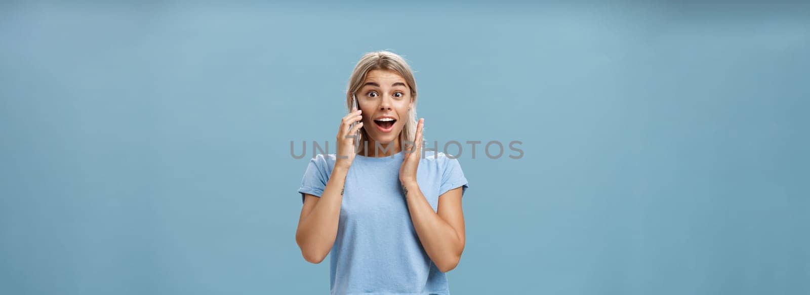 Pleased to hear best friend calling from abroad. Portrait of surprised pleased charming blonde female in trendy t-shirt opening mouth from amazement holding smartphone near ear and hand on face. Technology concept