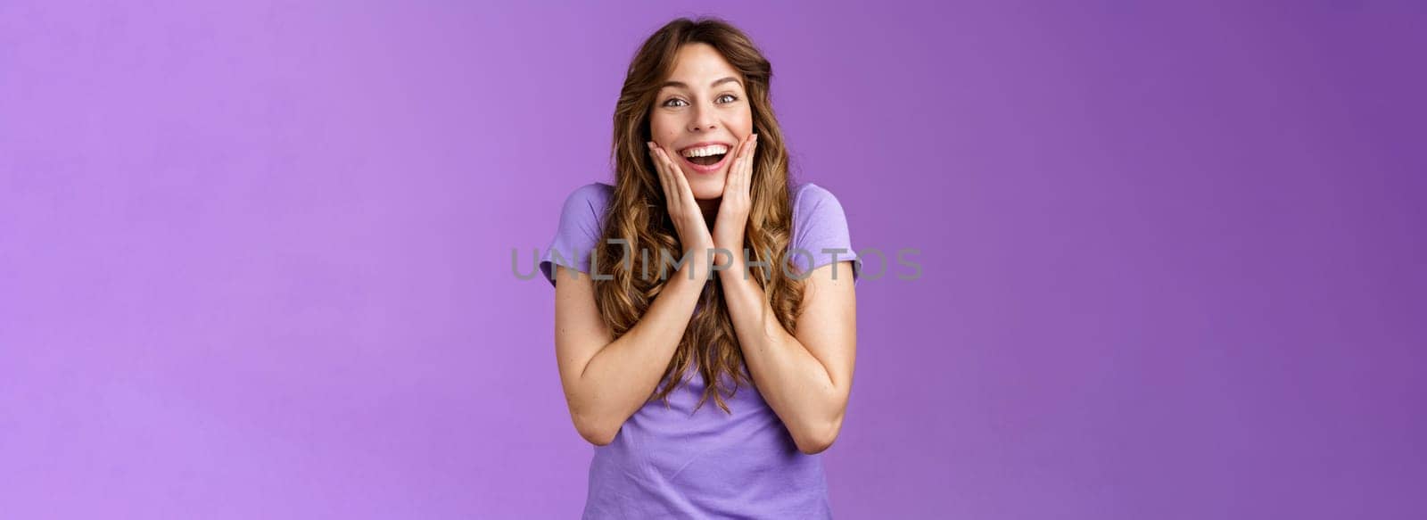Happy lively lucky enthusiastic girl curly hairstyle open mouth admiration fascinated smiling broadly touch cheek impressed surprised awesome incredible positive news stand purple background.