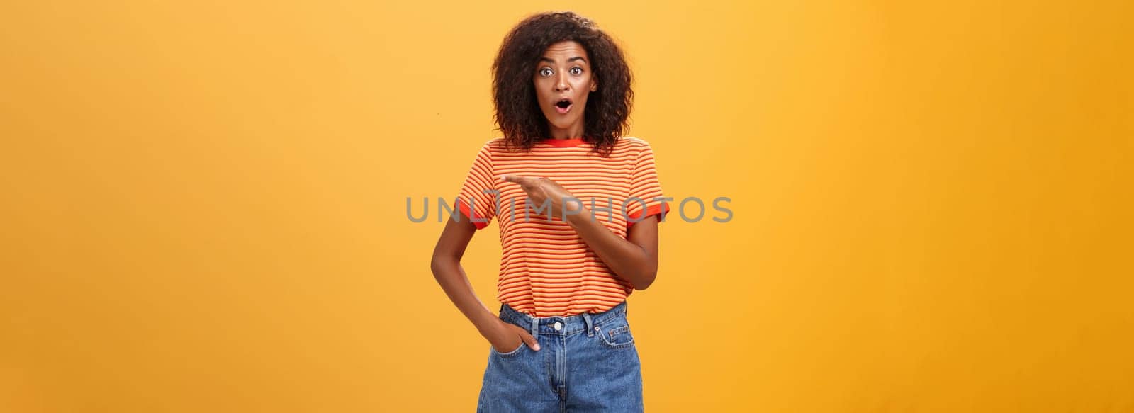 Studio shot of stylish fit female model in denim shorts and t-shirt gasping impressed pointing right while asking question being interested during observing masterclass over orange background. Lifestyle.