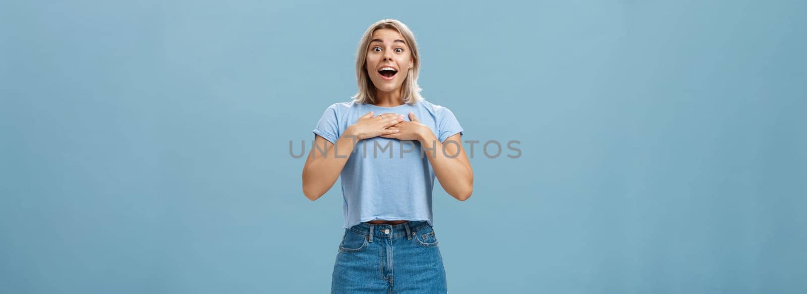 Lifestyle. Portrait of amazed and charmed attractive blond female student with tanned skin in denim shorts and summer t-shirt holding palms on breast gasping and smiling joyfully being grateful and pleased.