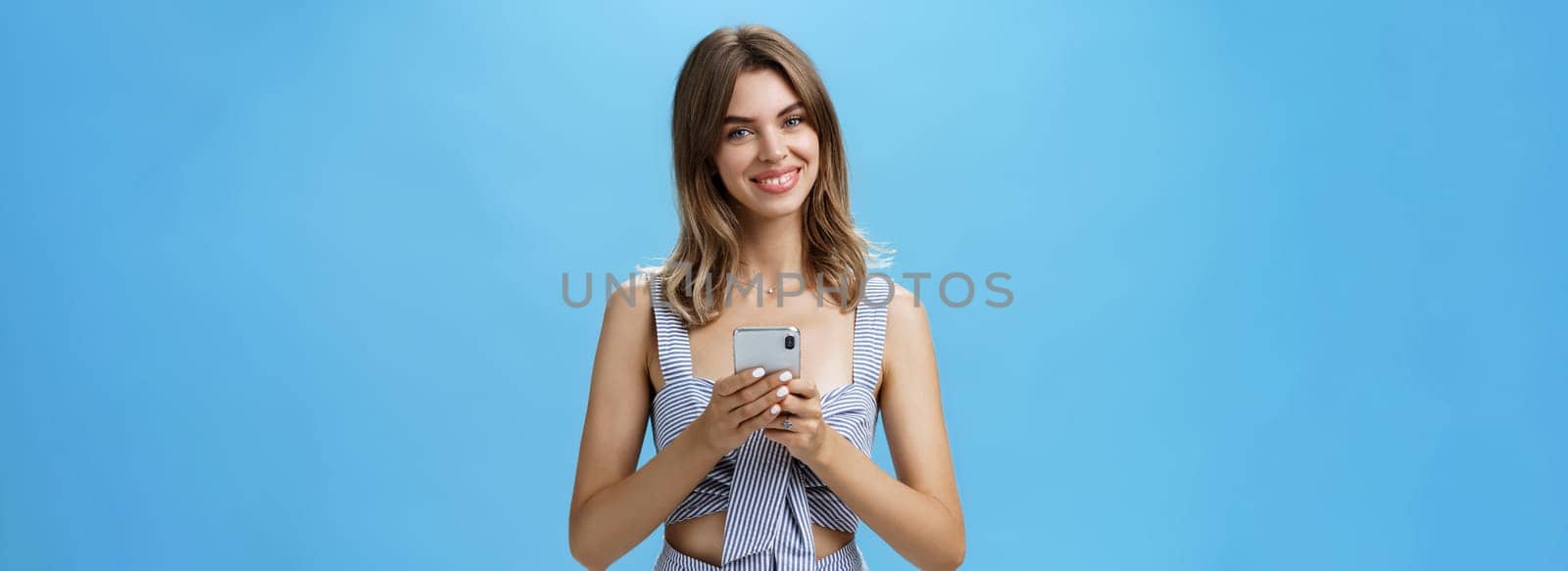Friendly pleasant good-looking woman in matching outfit holding smartphone over chest tilting head smiling broadly showing cute gapped teeth being delighted after reading heartwarming message by Benzoix
