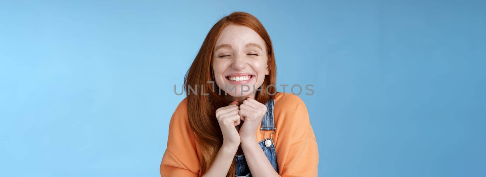 Eager rejoicing thrilled pretty young redhead girl close eyes dreamy smiling receive great result scholarchip triumphing joyfully grinning squeez hands excited, standing blue background very happy.