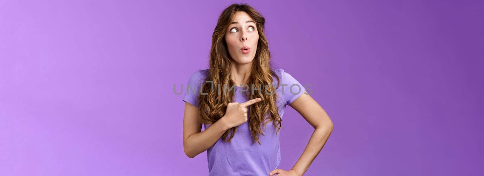Intrigued lively attractive feminine curly-haired female pointing looking left impressed folding lips say wow astonished express admiration enthusiastic check out awesome promo purple background by Benzoix