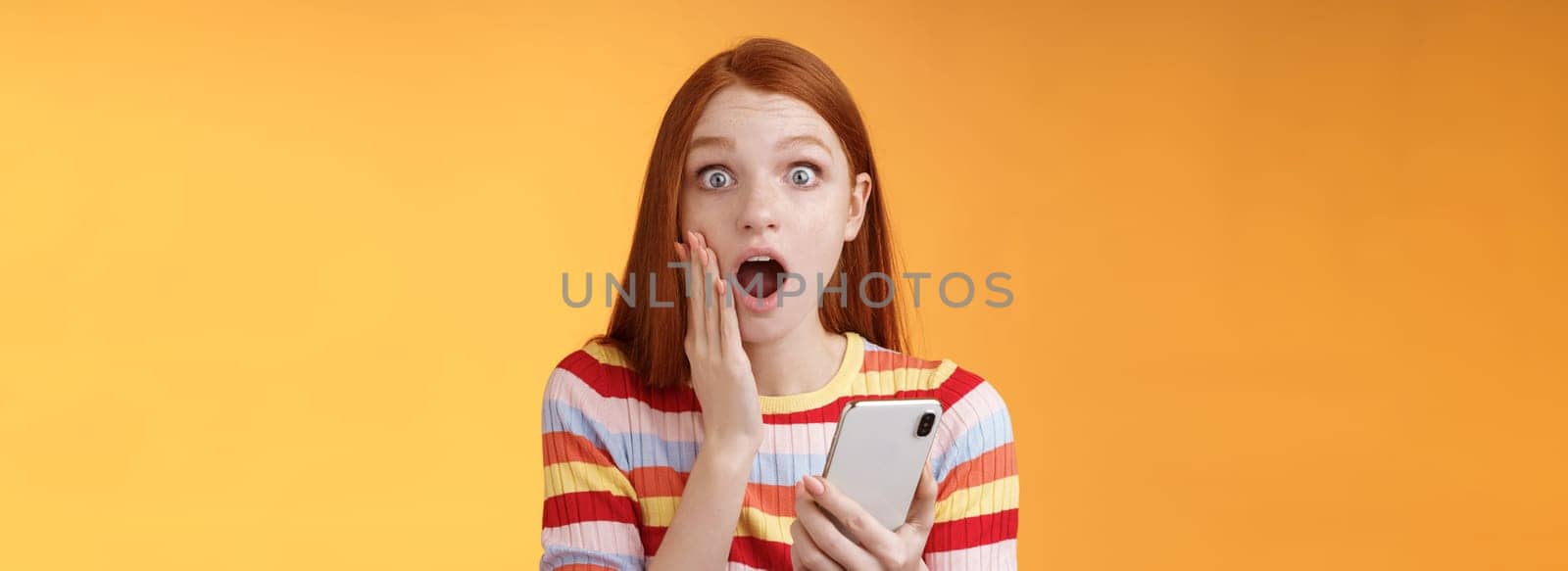 Concerned shocked emotive redhead girl find out stunning news look camera drop jaw gasping impressed touch cheek hold smartphone finish reading impressive disturbing story, orange background.