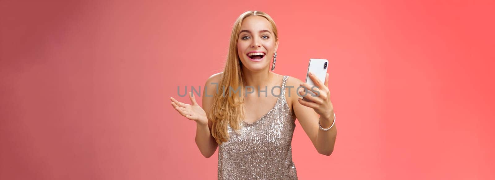 Astonished happy charming blond girl in silver glittering stylish dress holding smartphone amazed liking awesome result edit photo app smiling wondered amused, standing red background.