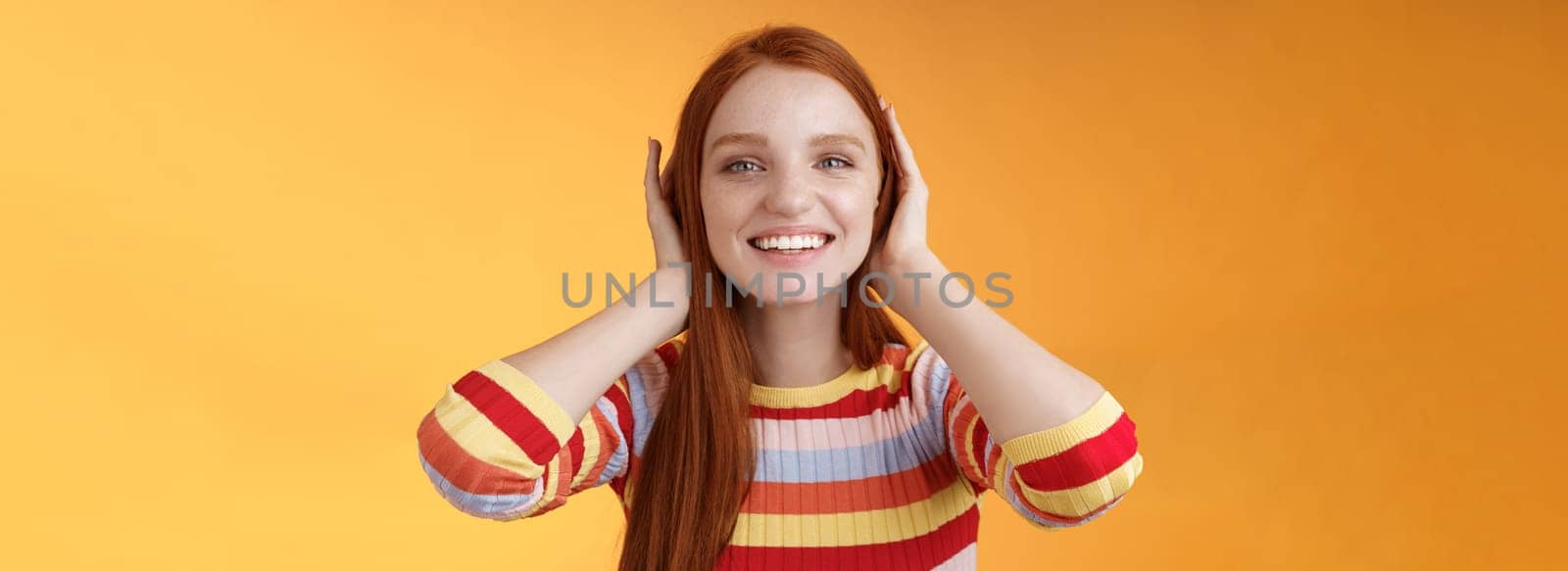 Cute playful tender smiling ginger girl blue eyes silly cover ears palms grinning fool around not hear anyone carefree unwilling listen noisy people standing careless relaxed orange background by Benzoix