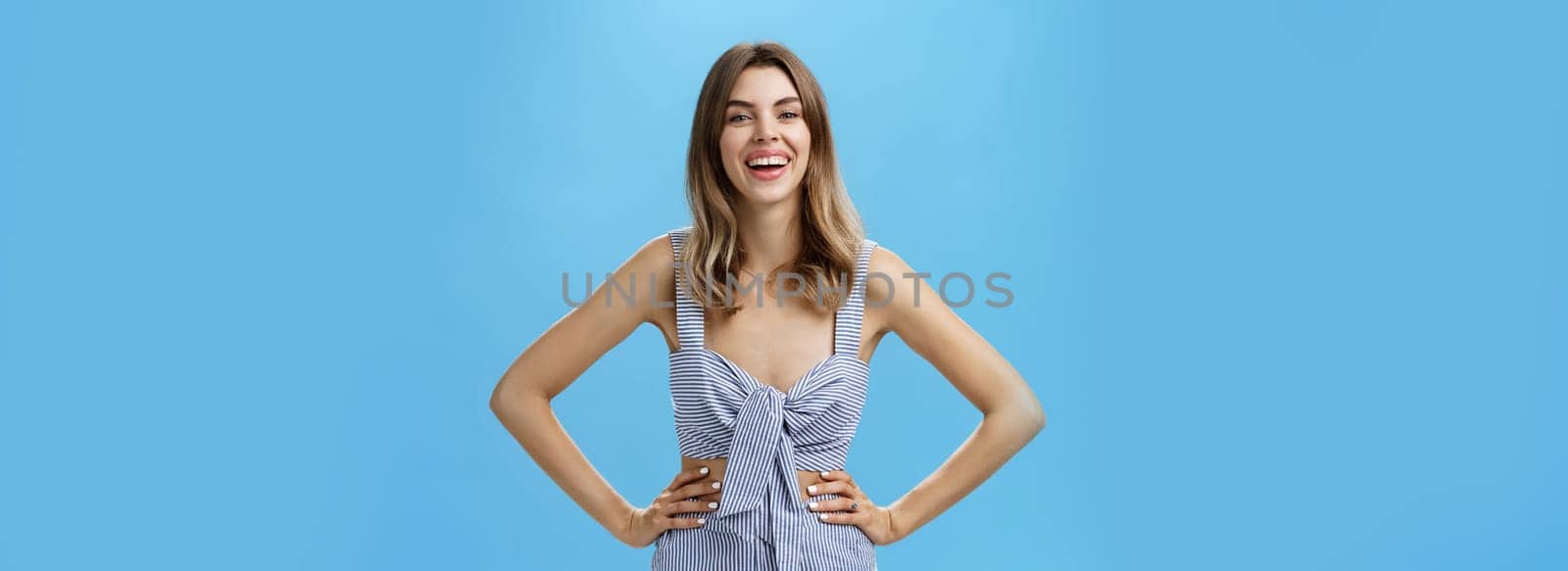 Amused and delighted cute adult female with chestnut hair in casual clothes holding hands on hip laughing and smiling broadly feeling excited during conversation posing against blue background. Copy space