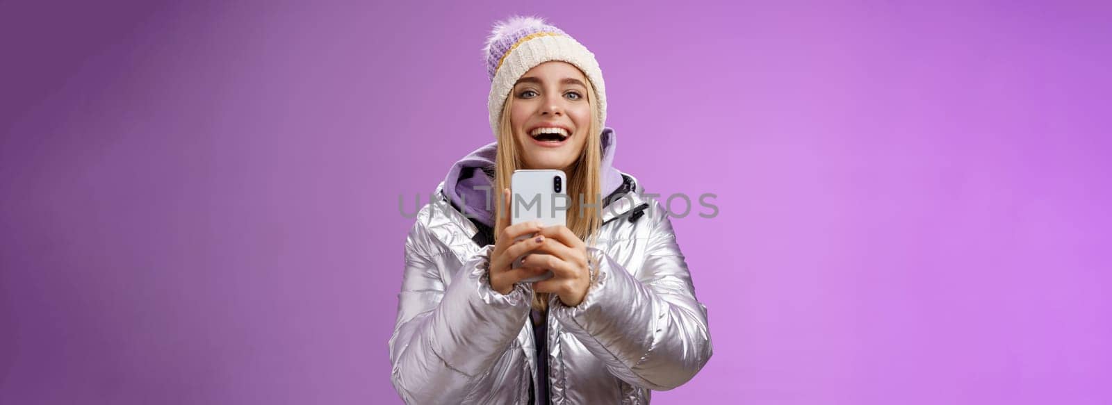 Amused excited attractive blond girlfriend holding smartphone up recording video boyfriend step snowboard first time capturing memories mobile camera standing happily purple background.