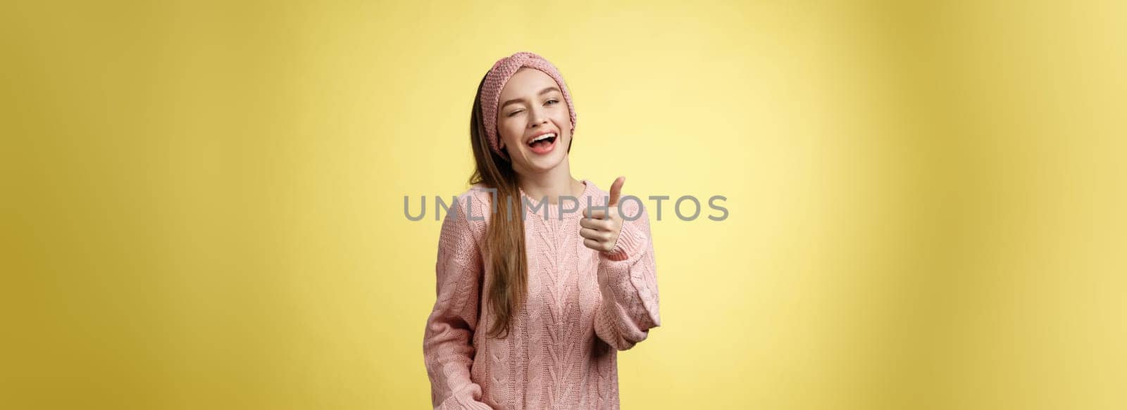 Happy amused beautiful young european girl in sweater, knitted headband showing good gesture, satisfied with service provided, winking sassy smiling with thumb-up in approval, giving recommendation.