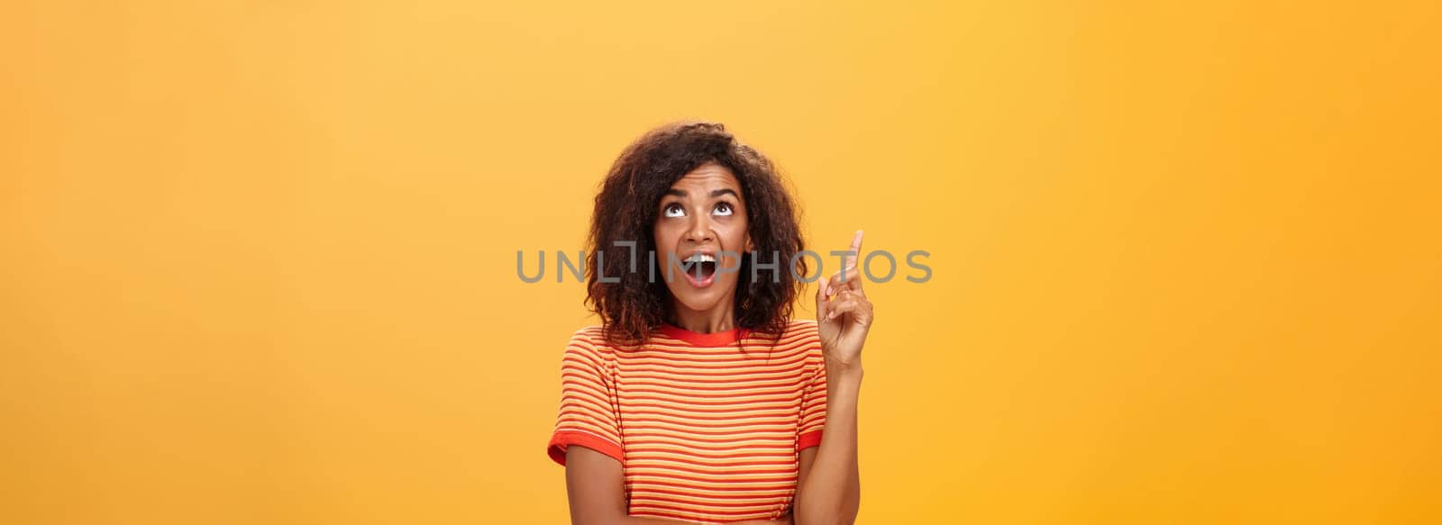 Waist-up shot of interested curious good-looking dark-skinned female in striped t-shirt talking asking question about curious star looking and pointing up with joy over orange background by Benzoix