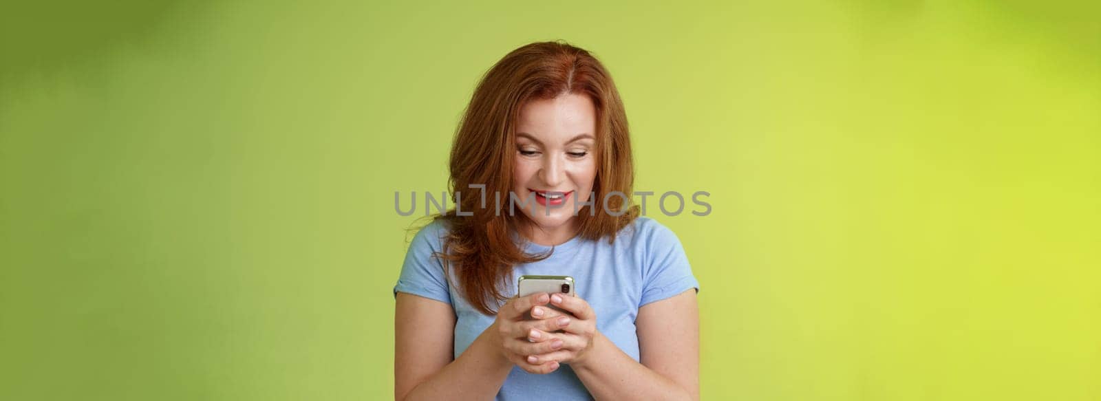 Intrigued excited tempting redhead middle-aged woman shopping online smartphone hold mobile phone look pleased entertained gadget screen smiling delighted playing game found perfect app by Benzoix