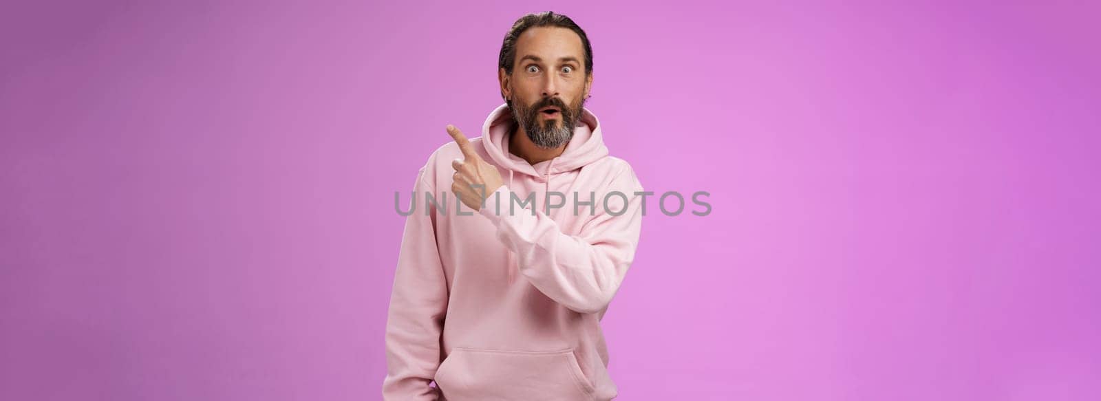 Amused handsome hipster mature 40s bearded caucasian man grey hair wearing trendy pink hoodie fold lips amazed wow pointing upper right corner widen eyes surprised check out awesome product by Benzoix