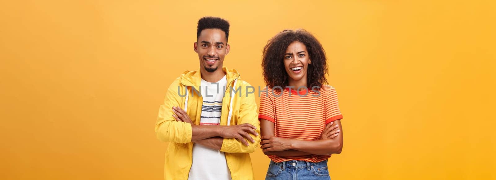 Portrait of surprised sarcastic african american woman with afro hairstyle standing with cute boyfriend crossing arms on chest laughing from scorn and fun over orange background. Copy space