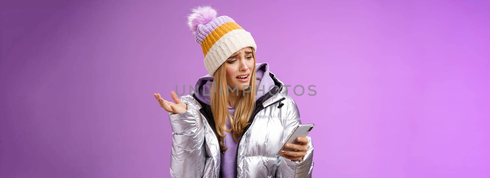 Upset disappointed attractive whining gloomy blond girl in silver jacket standing outside hat holding smartphone shrugging raising hand dismay complaining slow mobile internet, purple background.