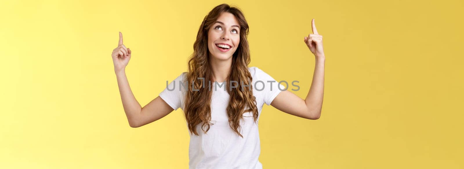 Happy admiring cute european curly-haired girl long haircut look pointing up impressed amused smiling broadly satisfactory delighted stand white t-shirt yellow background joyfully react good promo. Lifestyle.