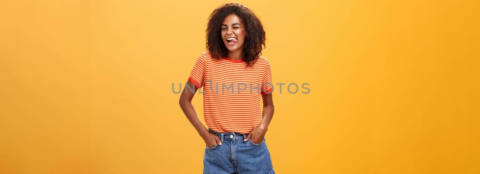 Woman sometimes wants feel childish. Joyful enthusiastic and charismatic young african american female with afro hairstyle winking happily showing tongue holding hands in pockets against orange wall.