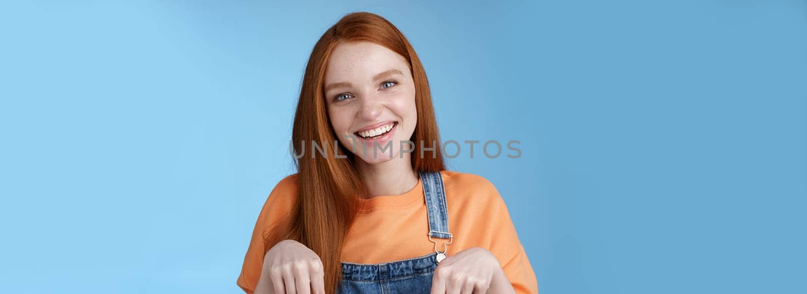 Friendly happy redhead girl smiling lively pretty grin pointing down index fingers offering good offer recommend use service standing blue background discuss interesting product, blue background.