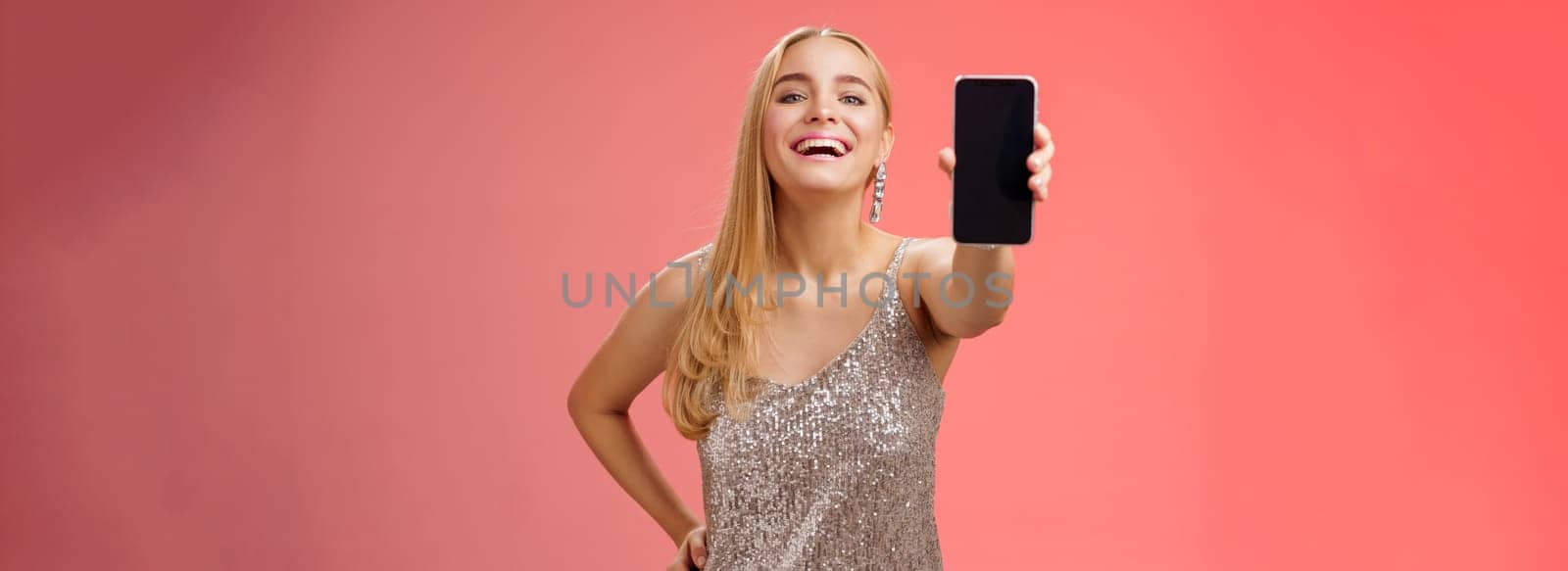 Proud joyful charming cheerful blond european woman in stylish silver shiny dress hold hand waist confident extend arm showing smartphone display present awesome new app device, red background.