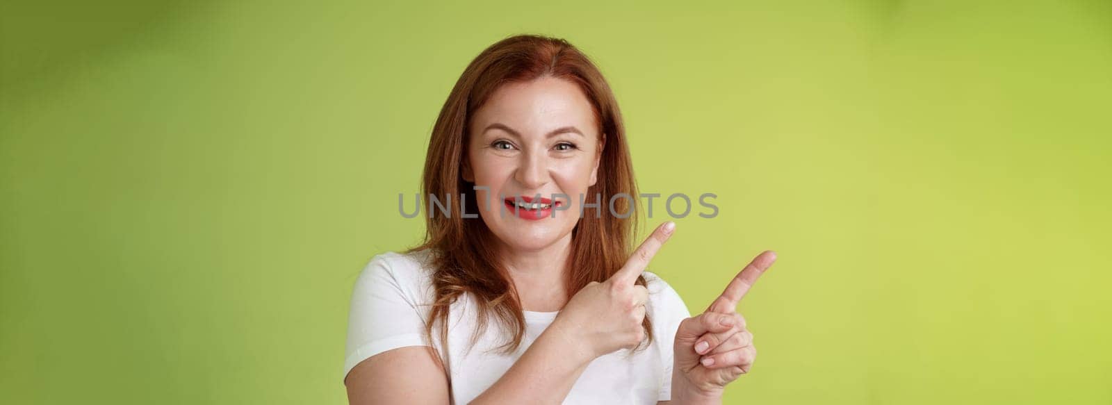 Close-up joyful motivated pleasant redhead middle-aged female. pointing upper left corner index fingers smiling delighted give advice check-out promo good advertisement blank space green background.