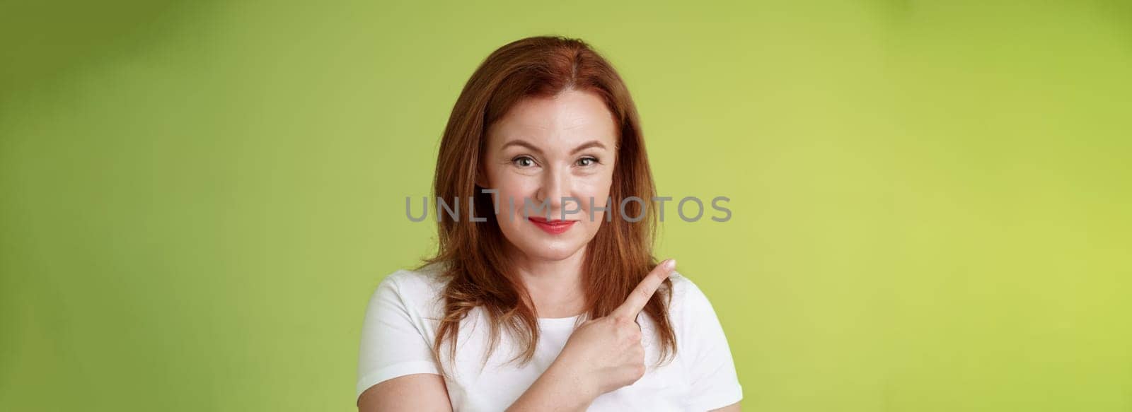 Kind cheerful good-looking middle-aged woman 50s redhead. white t-shirt smiling modest assured give advice pointing upper left corner indicating great promotion advertising product green background.