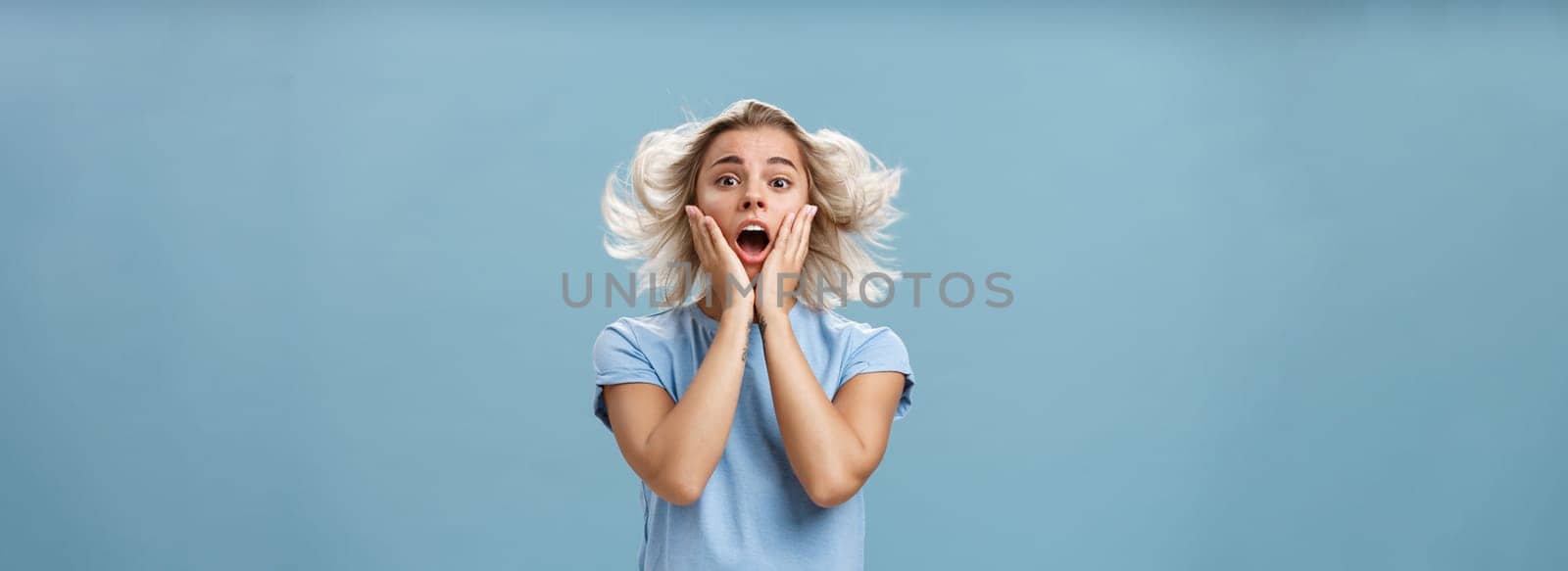 Indoor shot of shocked and impressed young stylish european female with blond hair holding hands on cheek from empathy and surprise opening mouth facing wind posing with hairstyle floating in air. Copy space