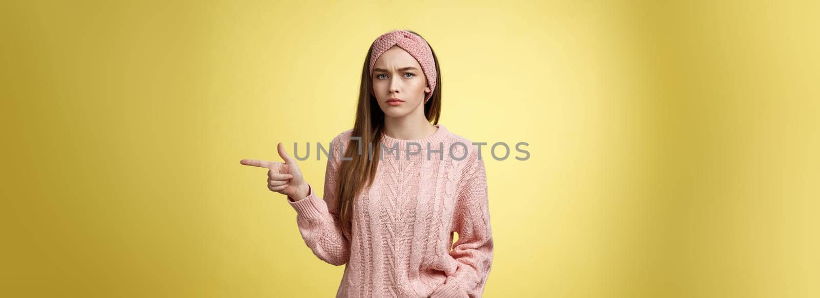 Pissed puzzled young attractive arrogant moody girlfriend in sweater, headband looking irritated, intense frowning pointing finger at copy space, directing person sits on her place, annoyed.