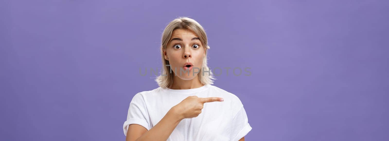 Waist-up shot of overwhelmed excited and surprised attractive urban female in white t-shirt folding lips gasping from amazement being questioned asking question and pointing left over purple wall by Benzoix