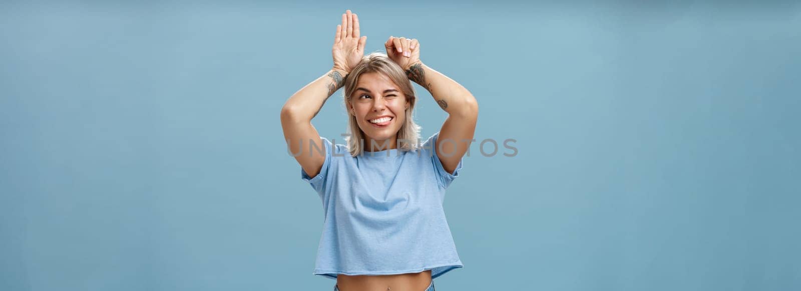 Studio shot of entertained carefree and emotive happy charming woman with tattoos on arms acting like bunny with palms on head winking joyfully smiling and sticking out tongue over blue wall by Benzoix